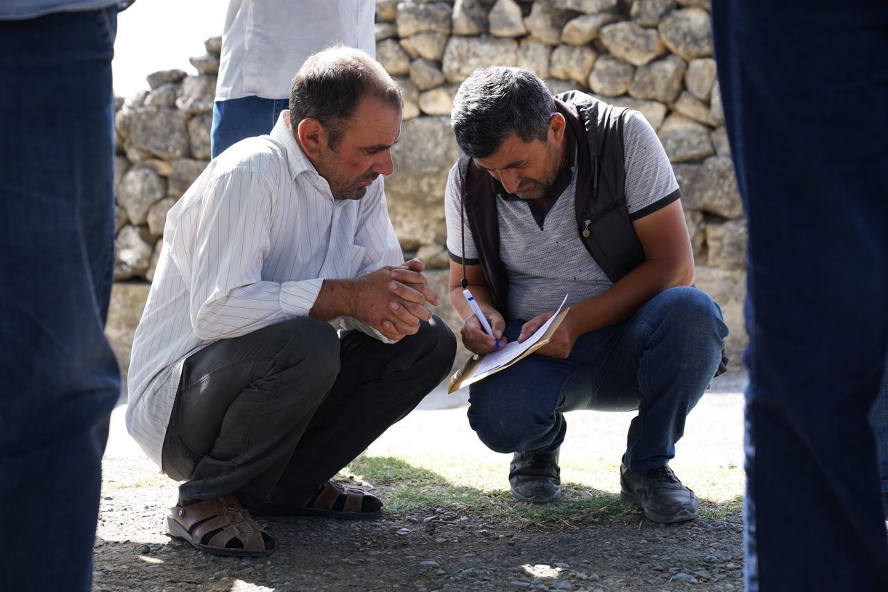 Two men squatting and analyzing paperwork outdoors in a sunny setting.